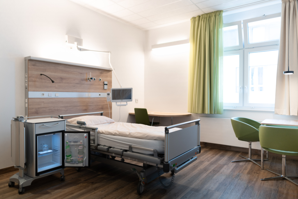 Patientenzimmer mit zwei Krankenhausbetten, Nachttischen und großen Fenstern für Tageslicht